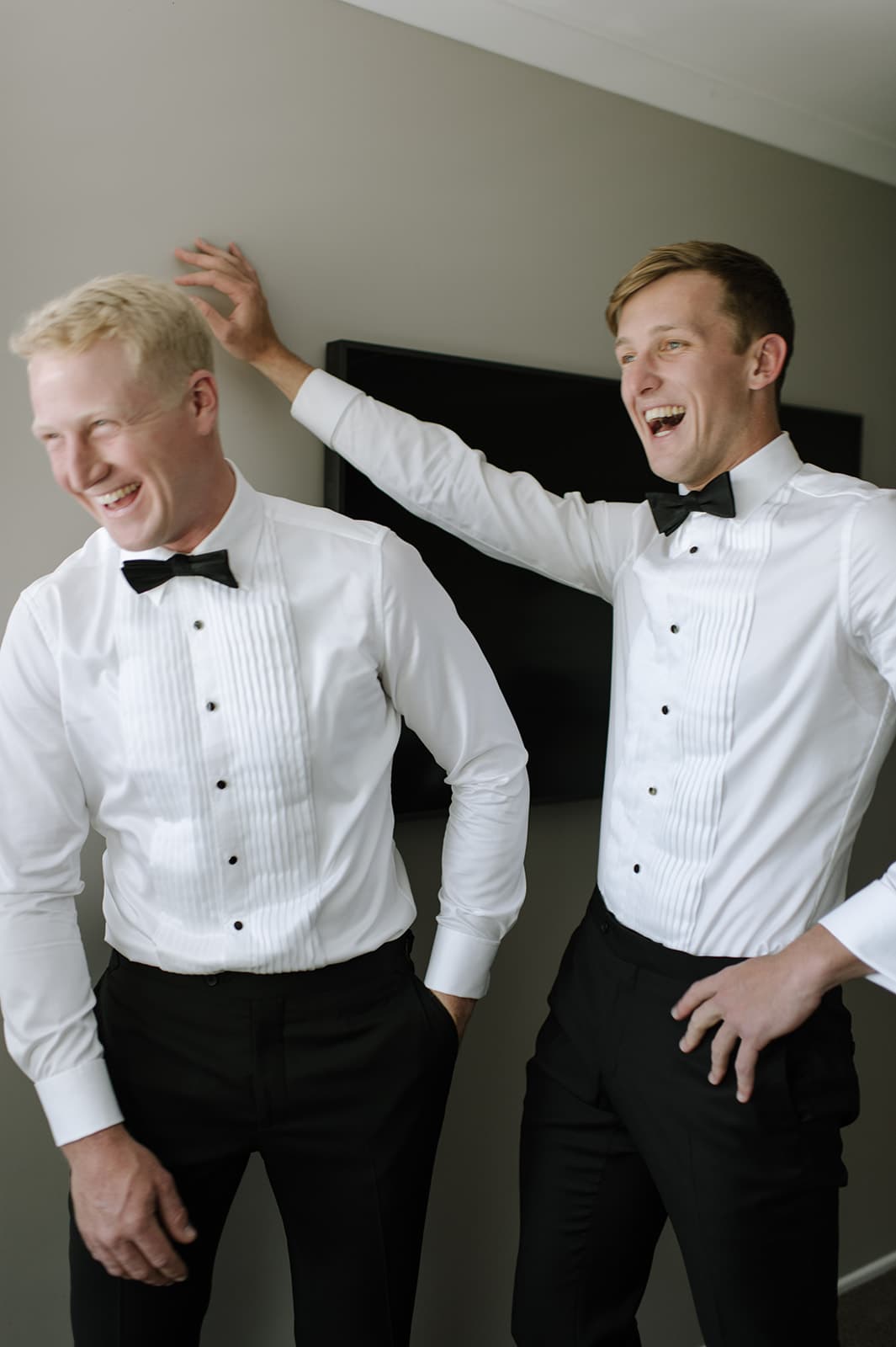 Two men are laughing and dressed in formal white shirts with black trousers and black bow ties. One has his hand on the other's shoulder. They are standing indoors against a muted wall. Both appear to be enjoying a light-hearted moment.