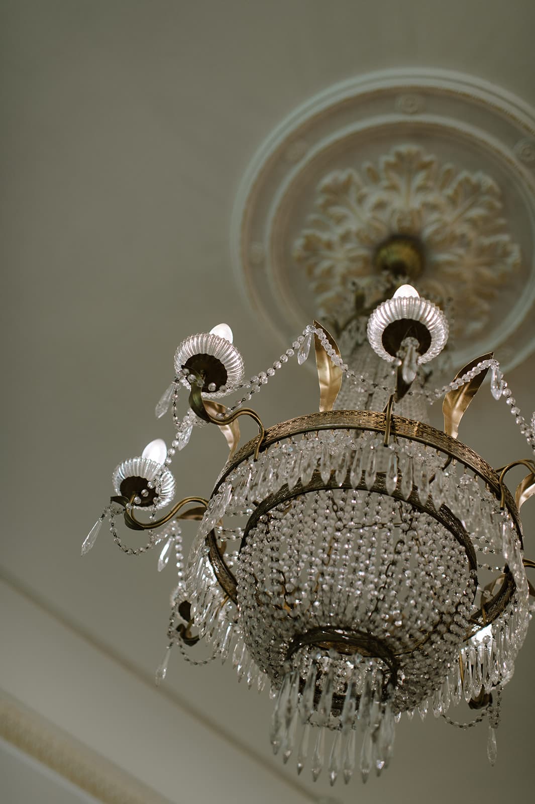 A close-up view of an ornate crystal chandelier with multiple tiers of hanging crystals, intricately designed metal arms, and candle-shaped light bulbs. The ceiling above features decorative molding. The chandelier is illuminated, casting a soft light.