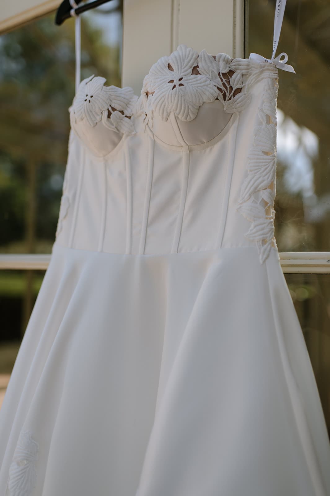 A close-up of a white strapless wedding dress hanging on a hanger. The dress has floral lace detailing around the bust and on the edges of the bodice. The fabric is smooth and the overall design of the dress is elegant and sophisticated.