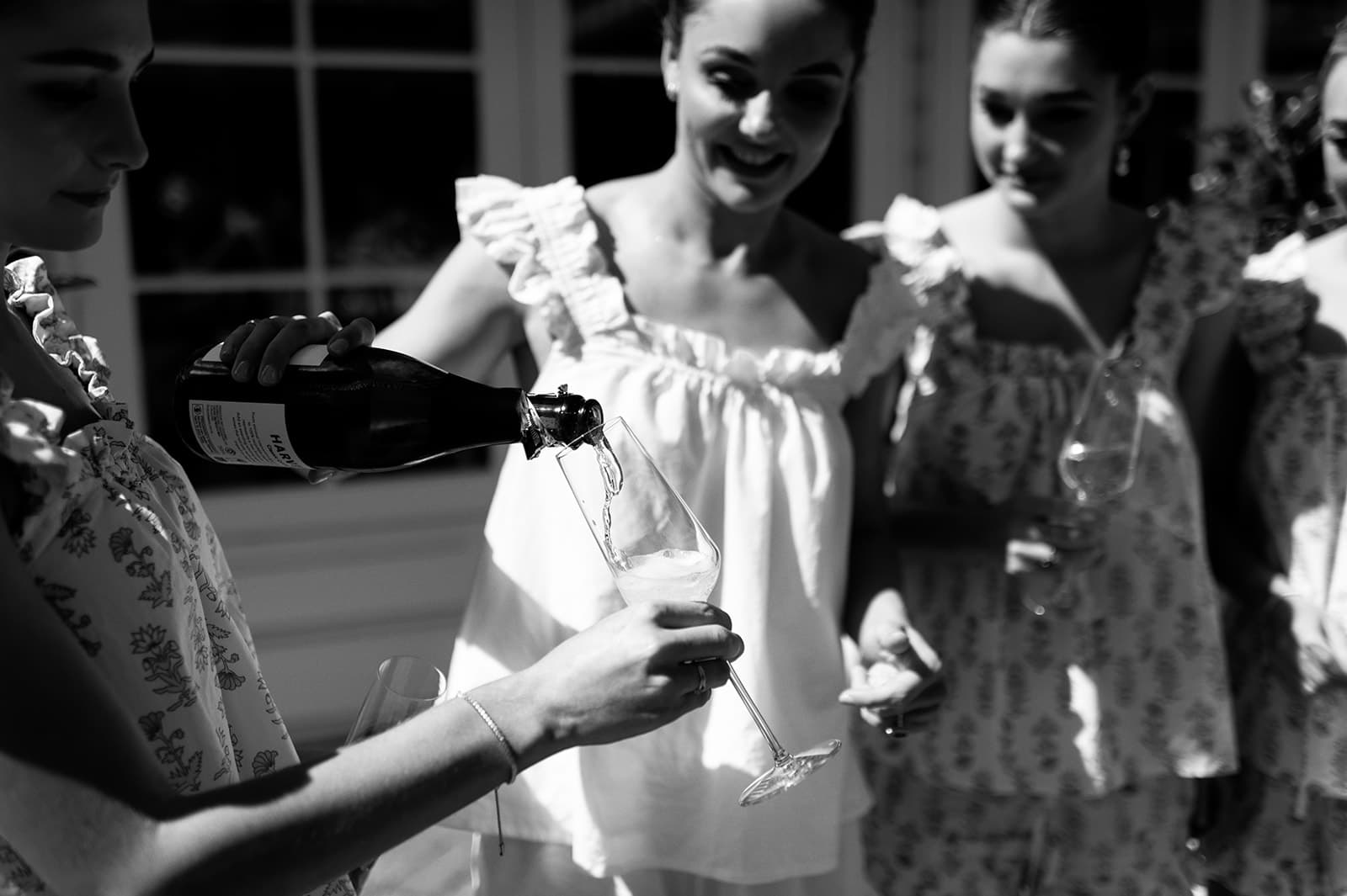 Four women wearing matching floral outfits enjoy a daytime gathering, with one pouring champagne into a glass held by another. The scene is in black and white, capturing their expressions of enjoyment and camaraderie against the backdrop of a well-lit room.