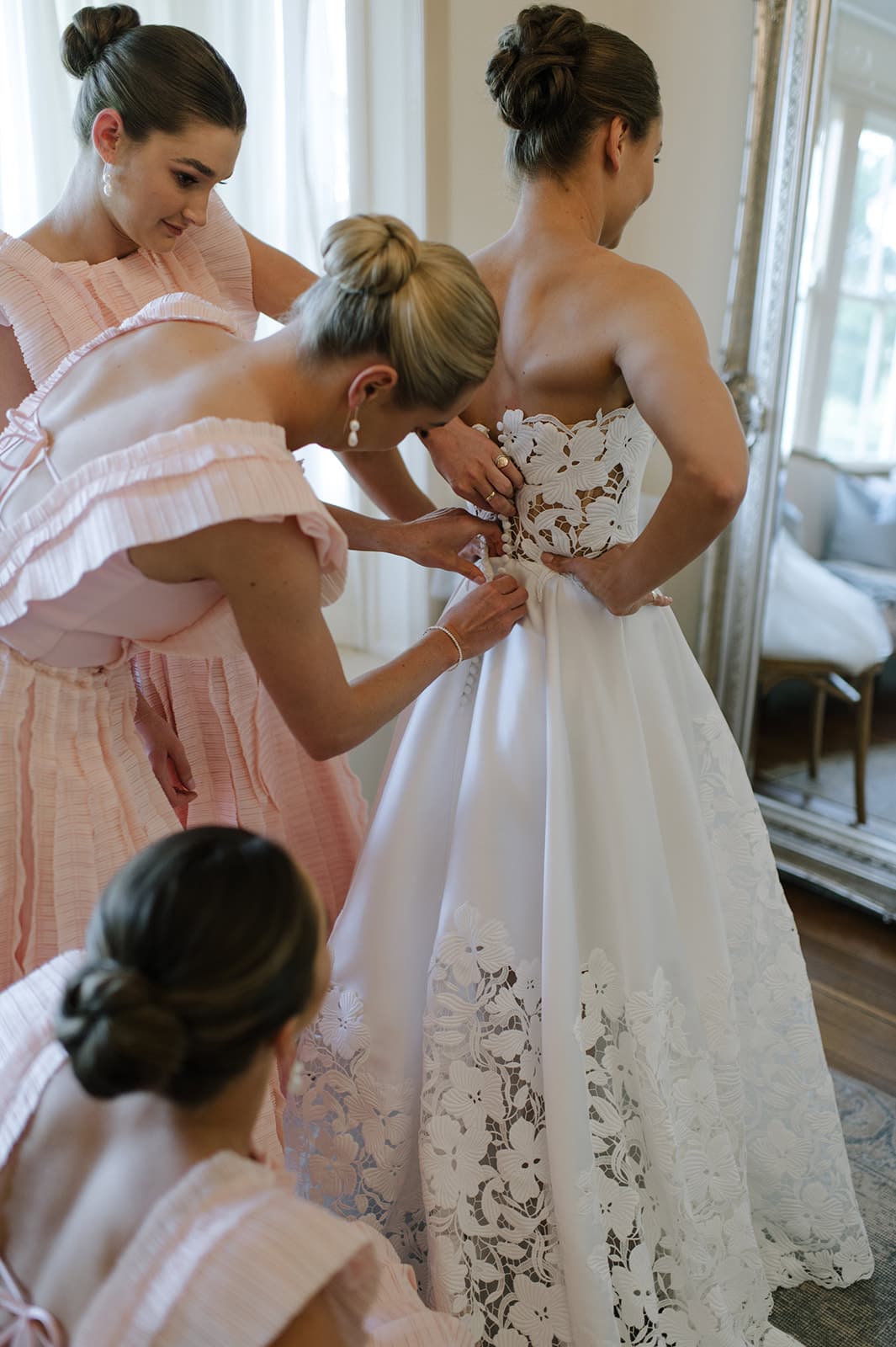 A bride in a white lace wedding gown is having her dress adjusted by three bridesmaids in matching pink dresses. They are in a well-lit room with large windows and a mirror. The bridesmaids are focused on ensuring the gown fits perfectly.