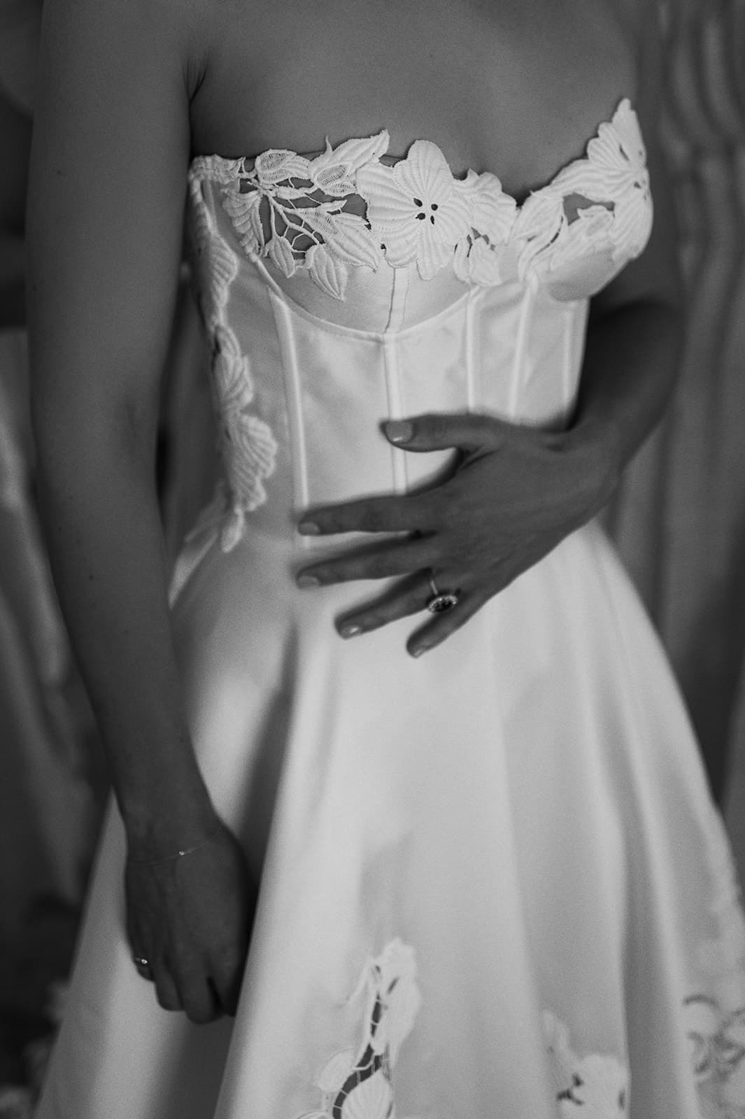 A black-and-white photo of a person in a strapless wedding dress with floral lace detailing. They are shown from the neck to the waist, holding their left hand, which displays a ring, gently against their stomach.