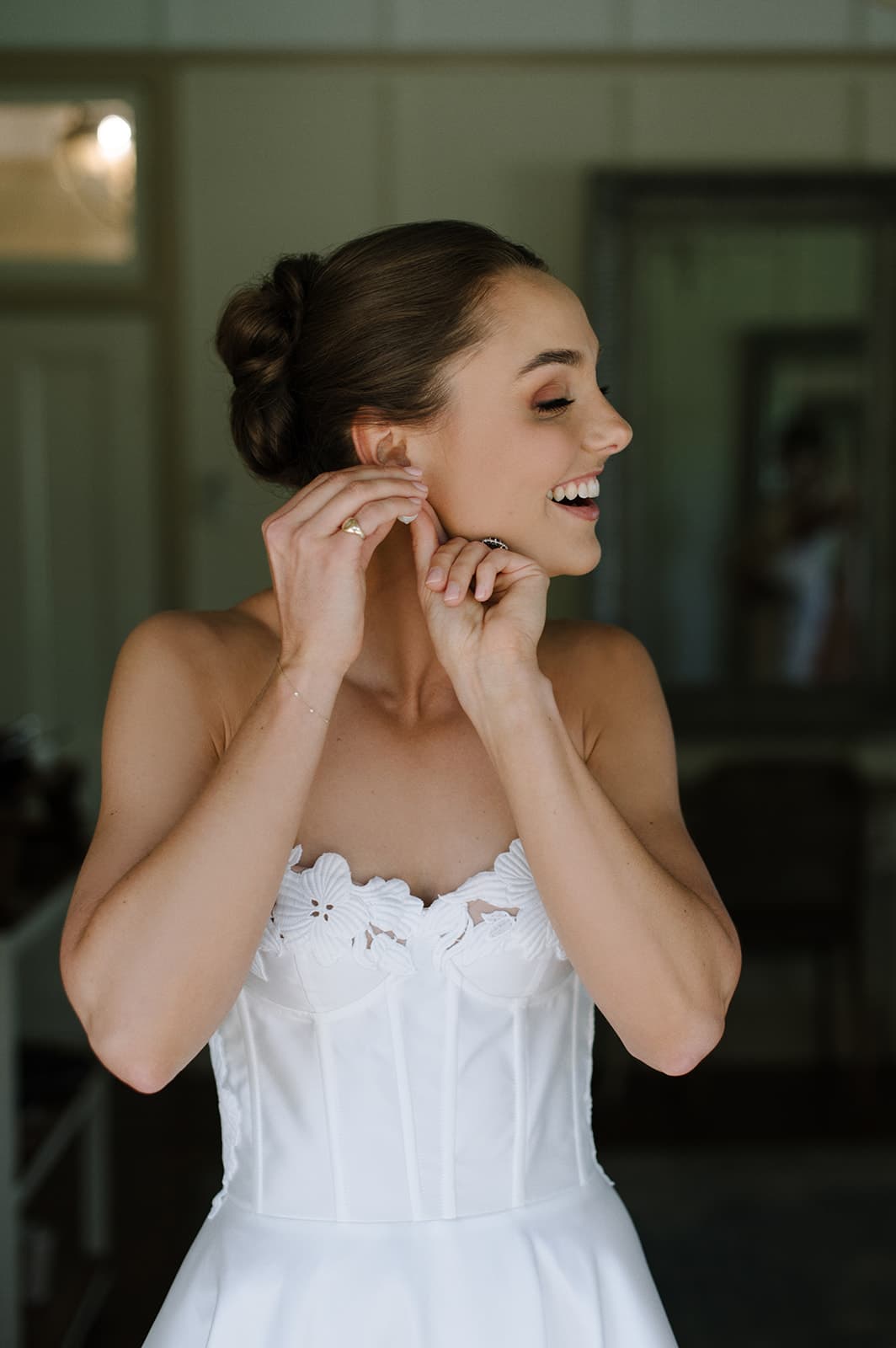 A bride with her hair in an elegant bun is smiling while putting on earrings. She is wearing a strapless white wedding dress featuring intricate lace detailing. She stands in a softly lit room with a mirror and furniture blurred in the background.