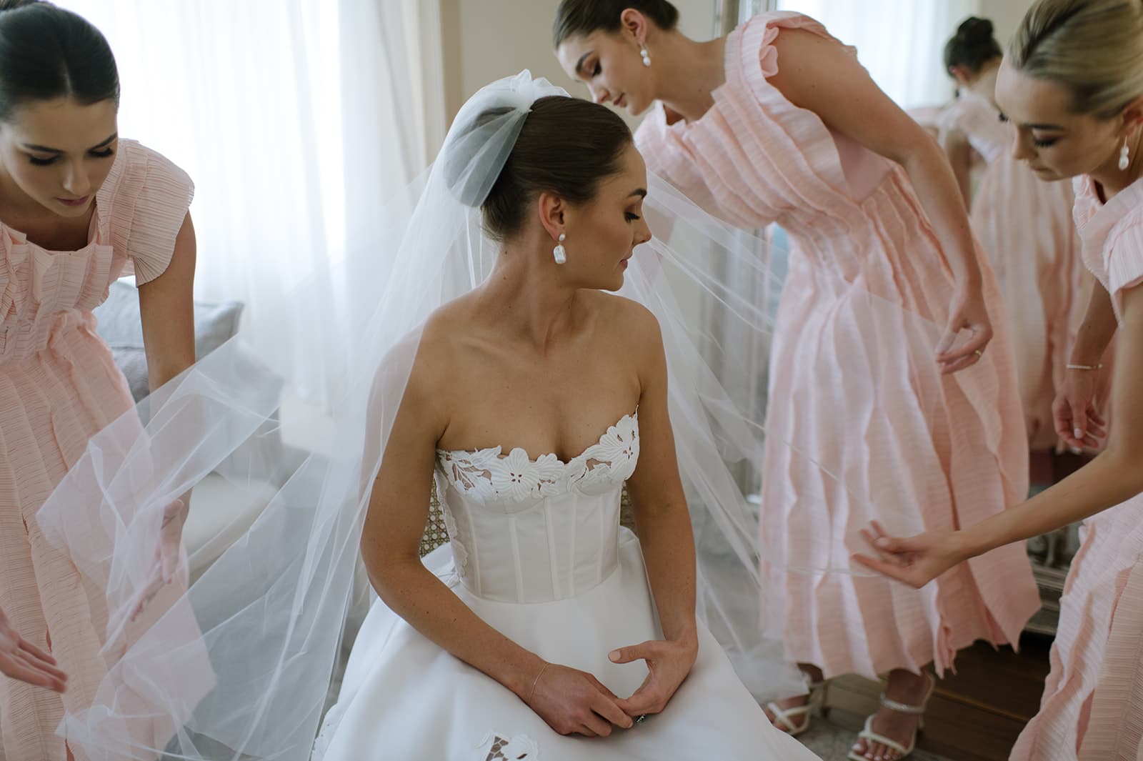 A bride, sitting down, is adjusting her white strapless wedding gown while wearing a veil. Three bridesmaids in matching light pink dresses assist her, each focusing on different parts of her veil and dress. The room has bright natural light coming through the window.