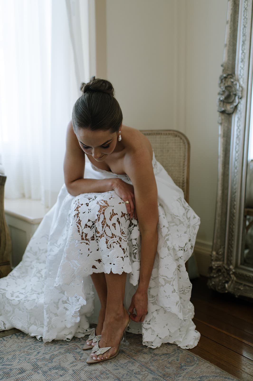 A bride in a white lace wedding dress sits on a wicker chair, bending down to adjust her shoe. She has her hair styled in an elegant updo and is in a softly lit room with a large window featuring sheer white curtains. An ornate large mirror stands next to her.