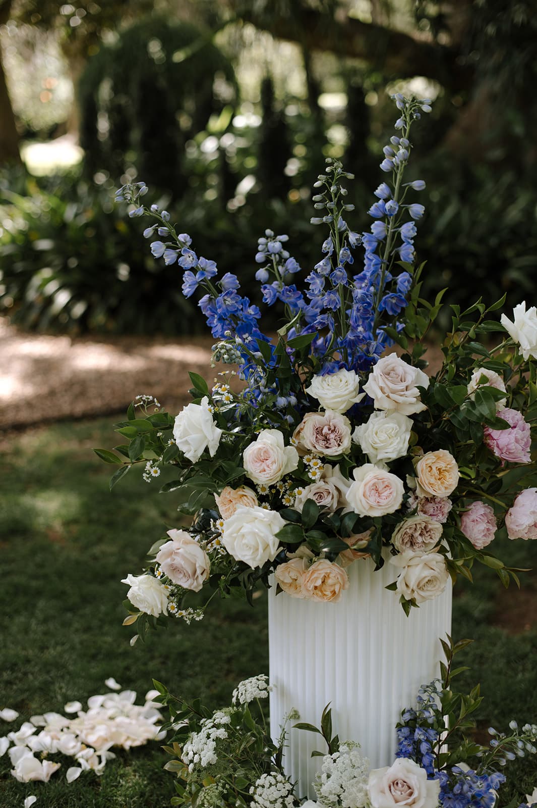 A tall, white, fluted vase holds a lush arrangement of roses in soft hues of white, pink, and peach, accented with sprigs of blue delphiniums and greenery. The vase is placed outdoors on green grass with a backdrop of lush plants and trees.