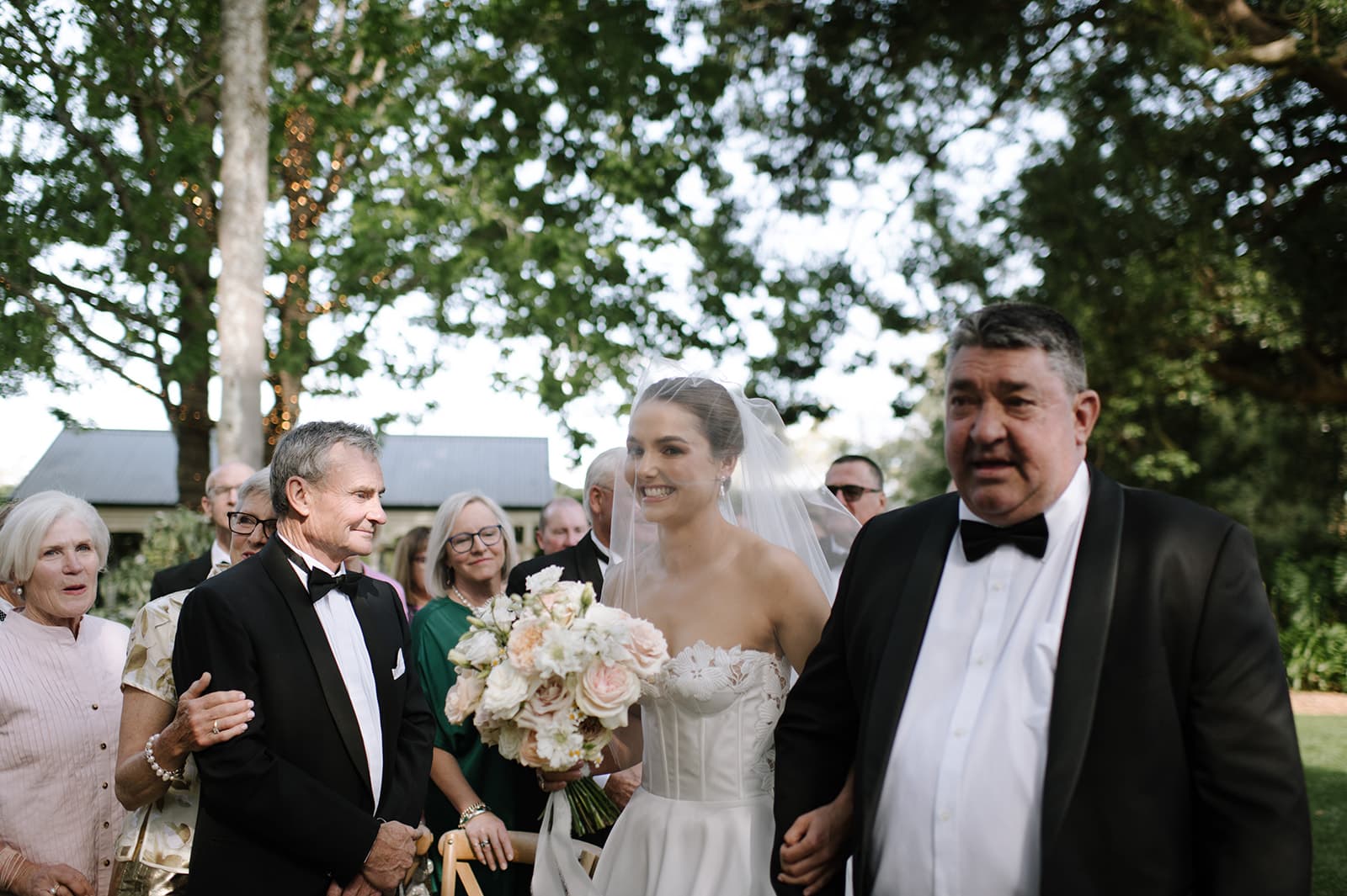 A bride in a white dress holding a bouquet walks down an outdoor aisle, accompanied by two men in tuxedos. Guests, some smiling and some looking emotional, stand on either side. Trees and greenery are visible in the background.