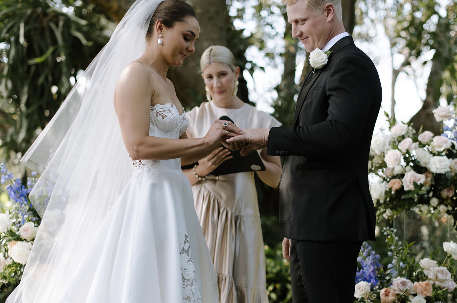 A bride and groom stand outdoors during their wedding ceremony. The bride, wearing a white strapless gown and veil, places a ring on the groom’s finger. The groom is in a black suit with a white rose boutonniere. A woman stands behind them holding a black folder.