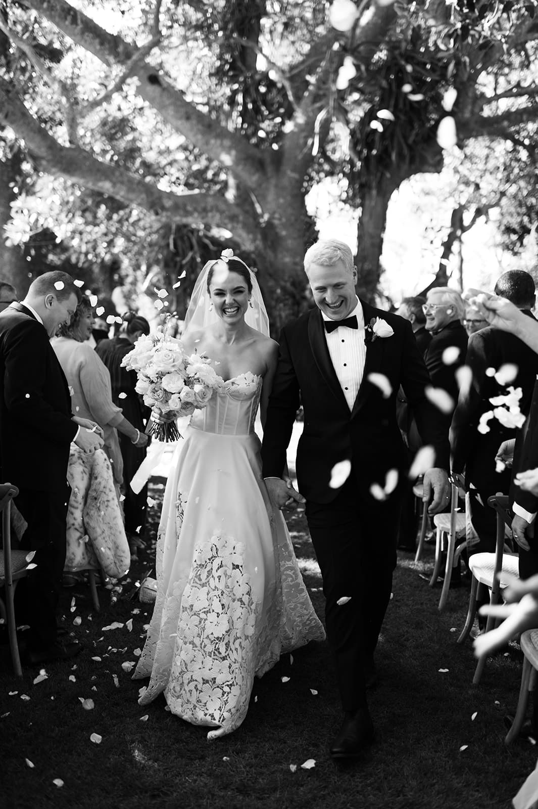 A bride and groom smile joyfully as they walk arm in arm down an outdoor aisle, surrounded by guests throwing flower petals. The bride wears a strapless gown and veil, holding a bouquet, while the groom is in a black suit. The scene is festive and celebratory.