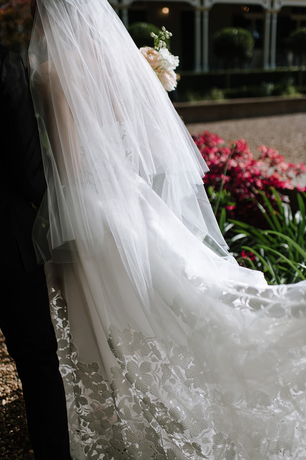 Close-up of a bride from the back in a white wedding dress adorned with intricate floral lace patterns. She is holding a bouquet of white flowers, and her long veil cascades elegantly down her back. A garden with blooming flowers is visible in the background.