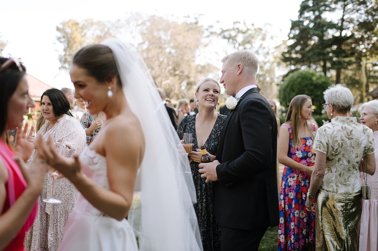 A bride in a white wedding dress talks to a guest in pink in the foreground. In the background, a couple, one in a black dress and the other in a black suit with a white rose boutonniere, smile while talking. Other guests are mingling in an outdoor garden setting.