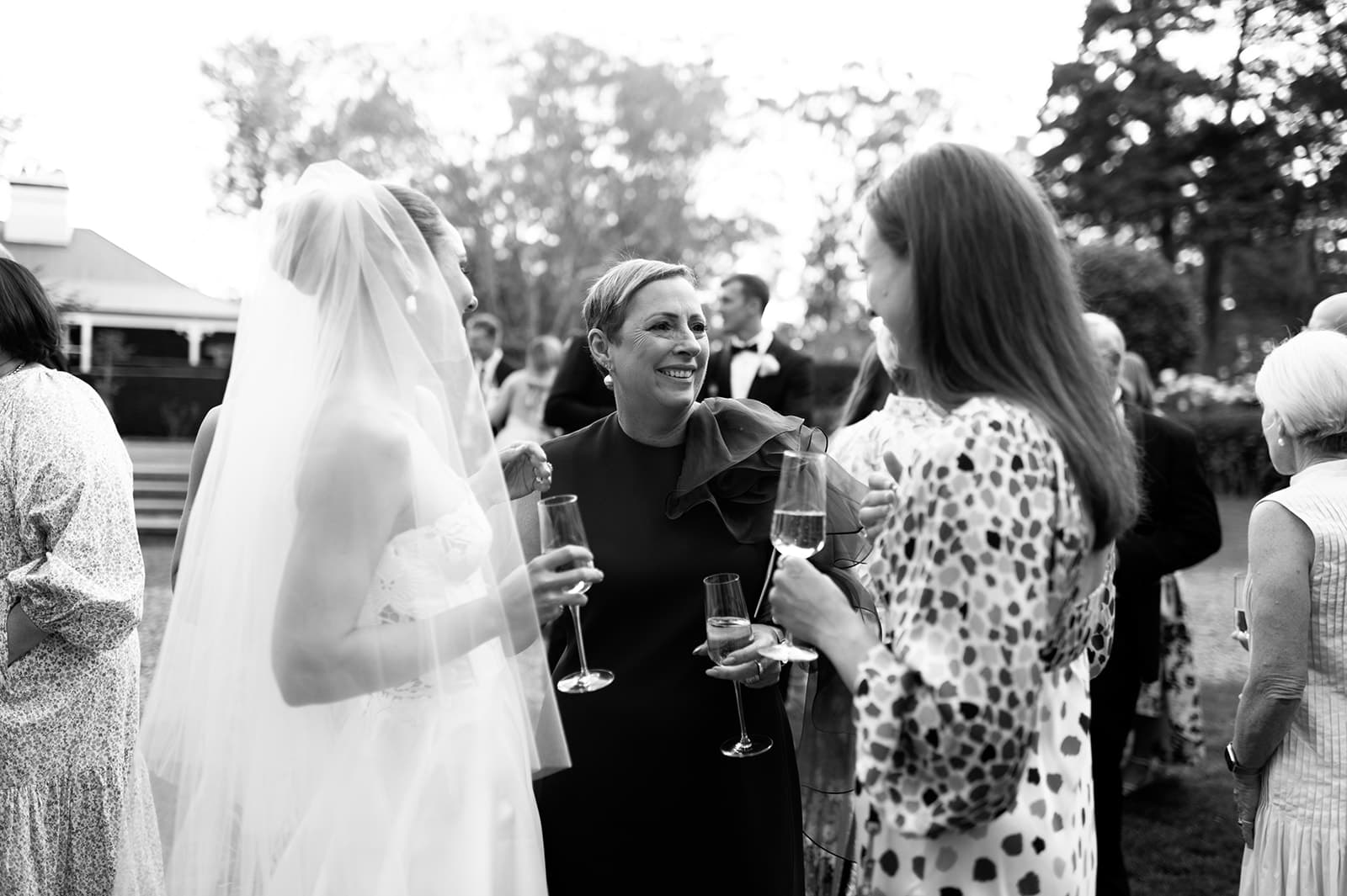 A black and white photo shows a bride in a wedding dress and veil holding a champagne flute, conversing and laughing with two women. They are outdoors, and other guests are seen socializing in the background. Trees and a house are partially visible.