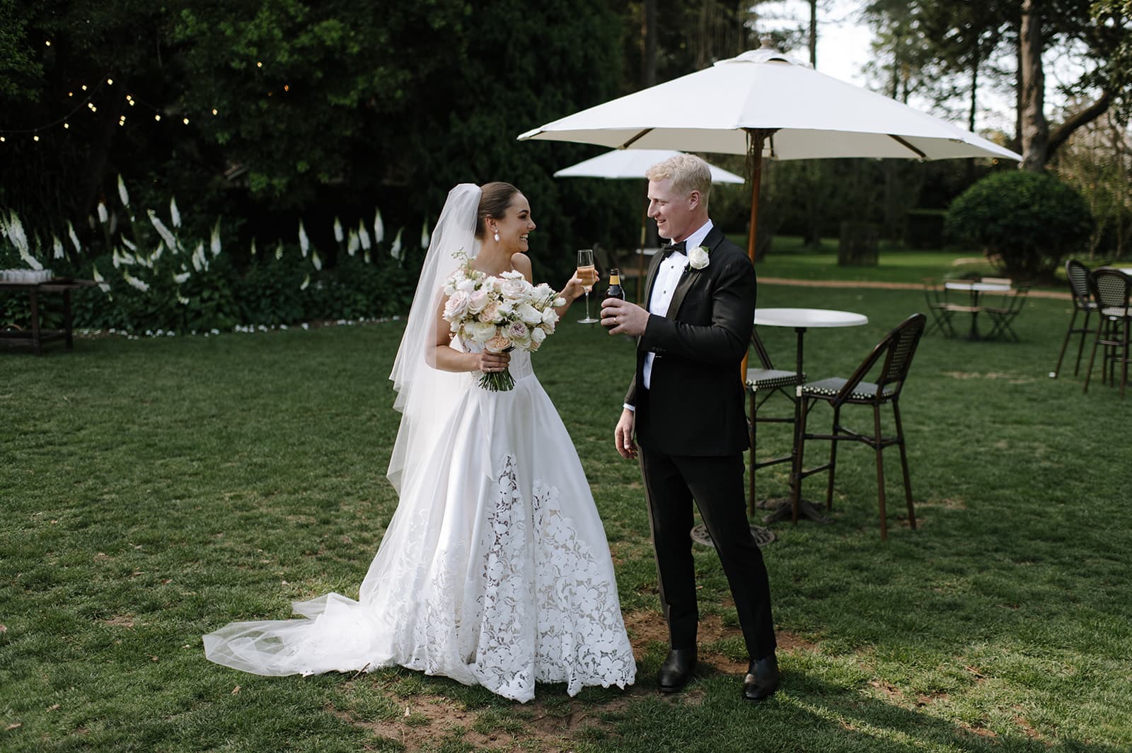 A bride in a white gown and veil stands beside a groom in a black tuxedo. They are outdoors on a lawn, under white umbrellas and string lights. The bride holds a bouquet of flowers, and they are clinking glasses. Greenery and trees surround them.