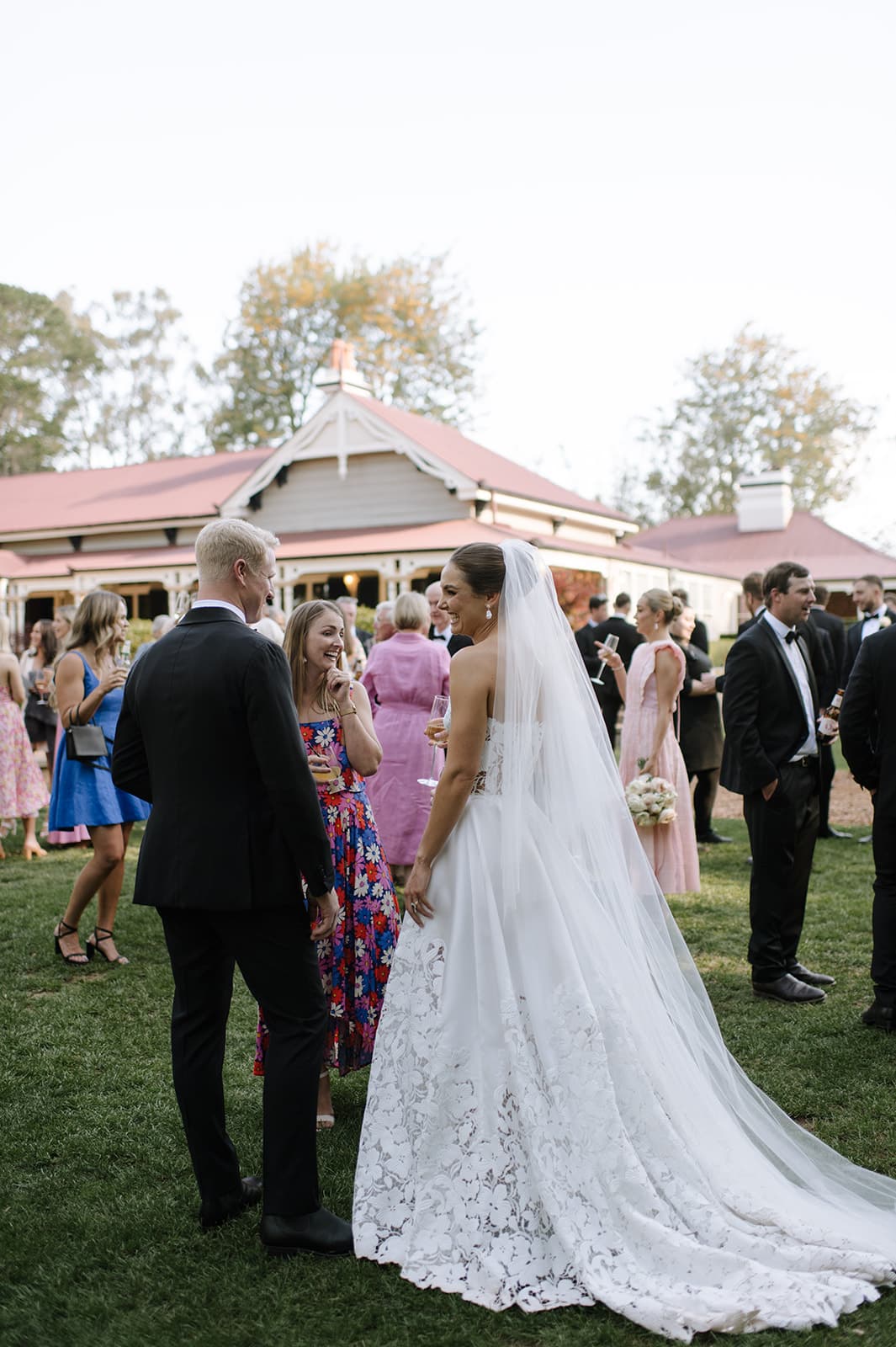 A bride in a white lace gown and veil stands outdoors, engaged in conversation with groom and guests. The groom is in a black suit. Guests dressed in formal attire are mingling in the garden of a house with a red roof, surrounded by trees on a sunny day.