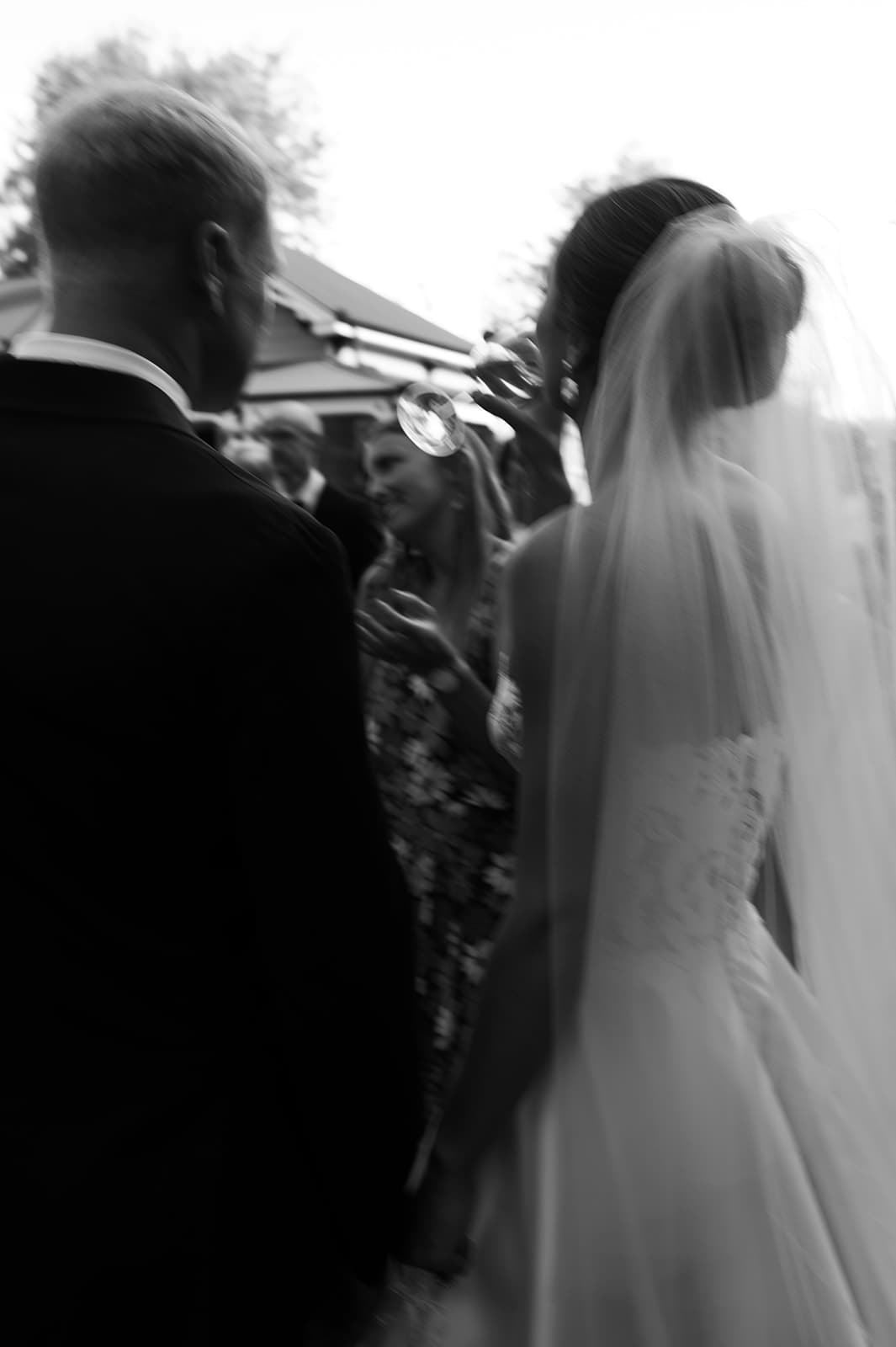 A black and white image showing a bride and groom from behind, walking hand in hand. The bride is wearing a long veil and a wedding gown, and the groom is in a suit. In the background, a group of guests can be seen, with one woman raising a glass.