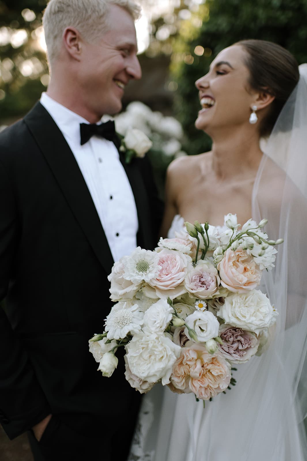 A groom in a black suit and bow tie and a bride in a white wedding dress share a joyful moment, smiling and looking at each other. The bride holds a large bouquet of light-colored flowers, and greenery is visible in the background.