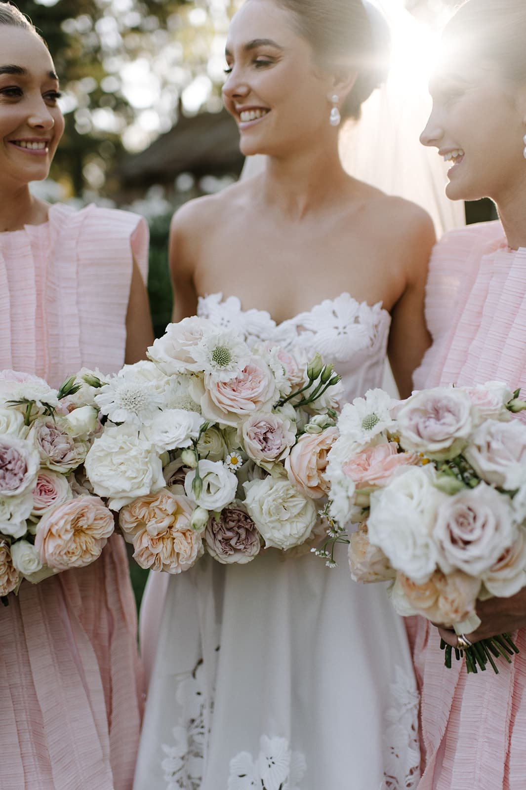 Three women, one in a white strapless wedding dress and veil, and two in light pink bridesmaid dresses, smile while holding bouquets of white and blush flowers. They stand close together, with sunlight filtering through the trees behind them.
