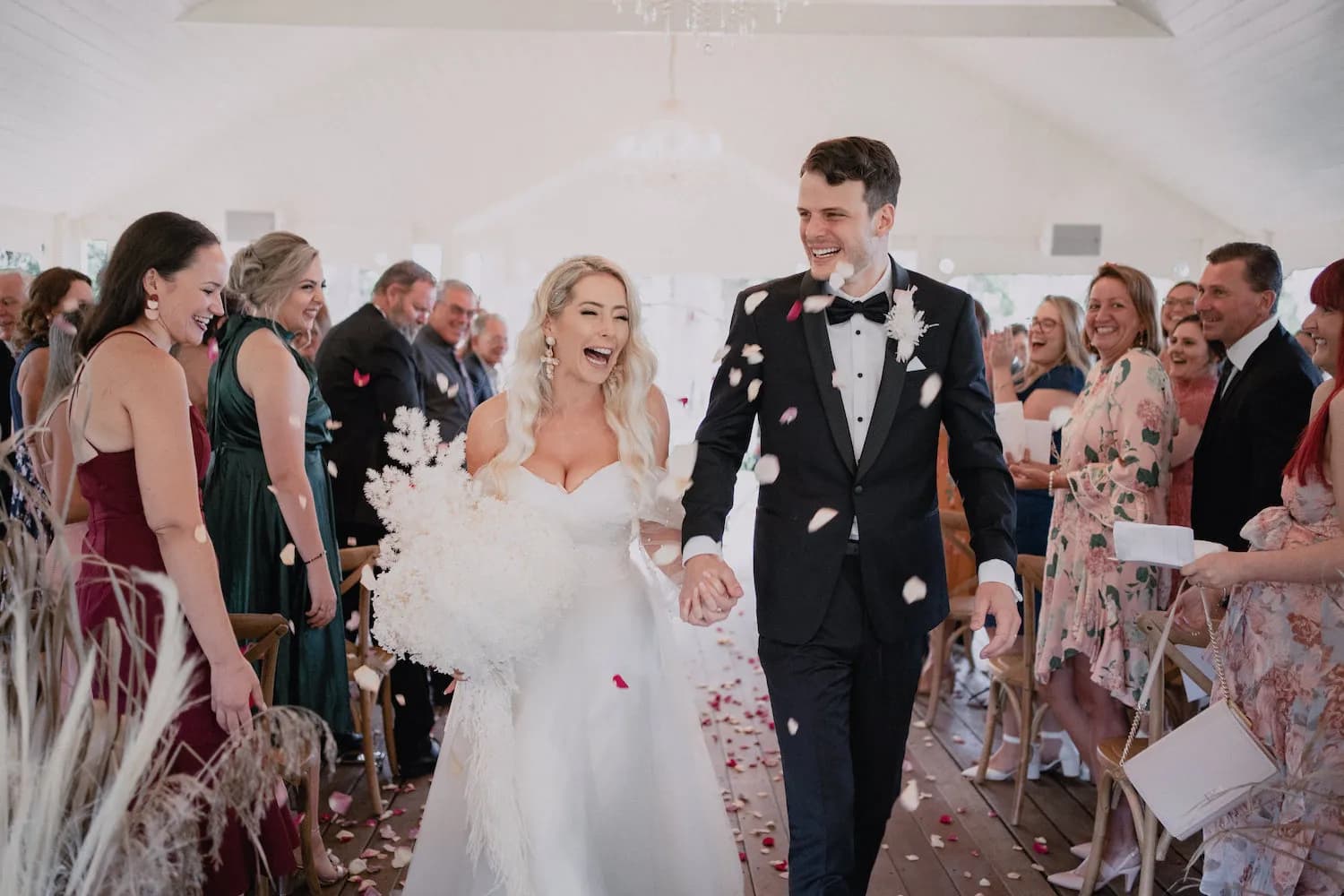 A joyful bride and groom walk down the aisle hand in hand, smiling and laughing as guests shower them with flower petals. The bride holds a bouquet of white flowers and wears a strapless white gown, while the groom is dressed in a black tuxedo.