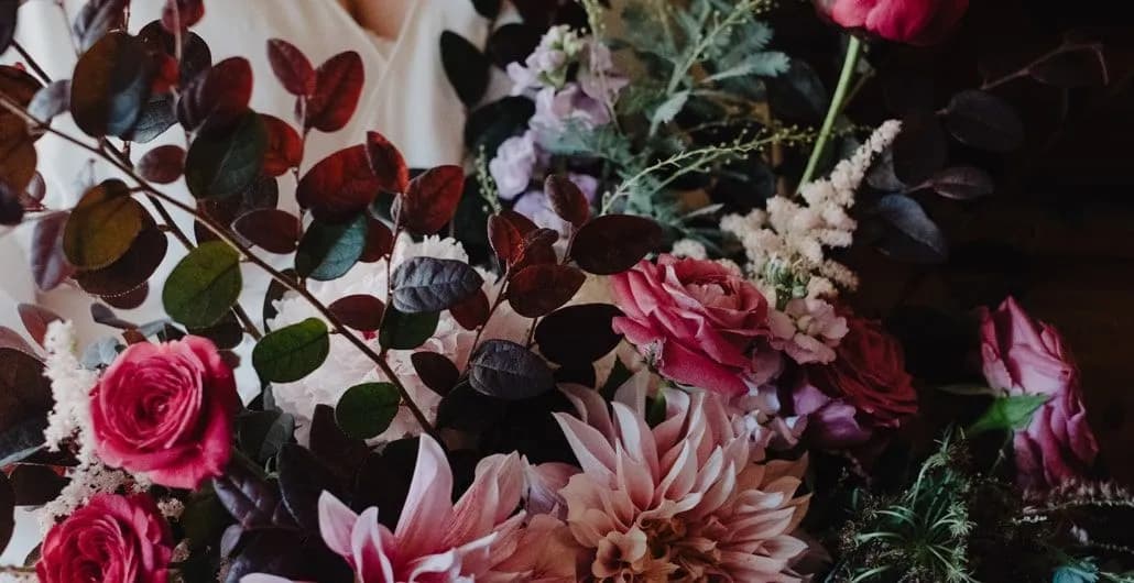 A bouquet comprised of various flowers and green foliage, including pink roses, white astilbe, and lush leaves. The background is blurred, highlighting the vibrant and intricate arrangement.