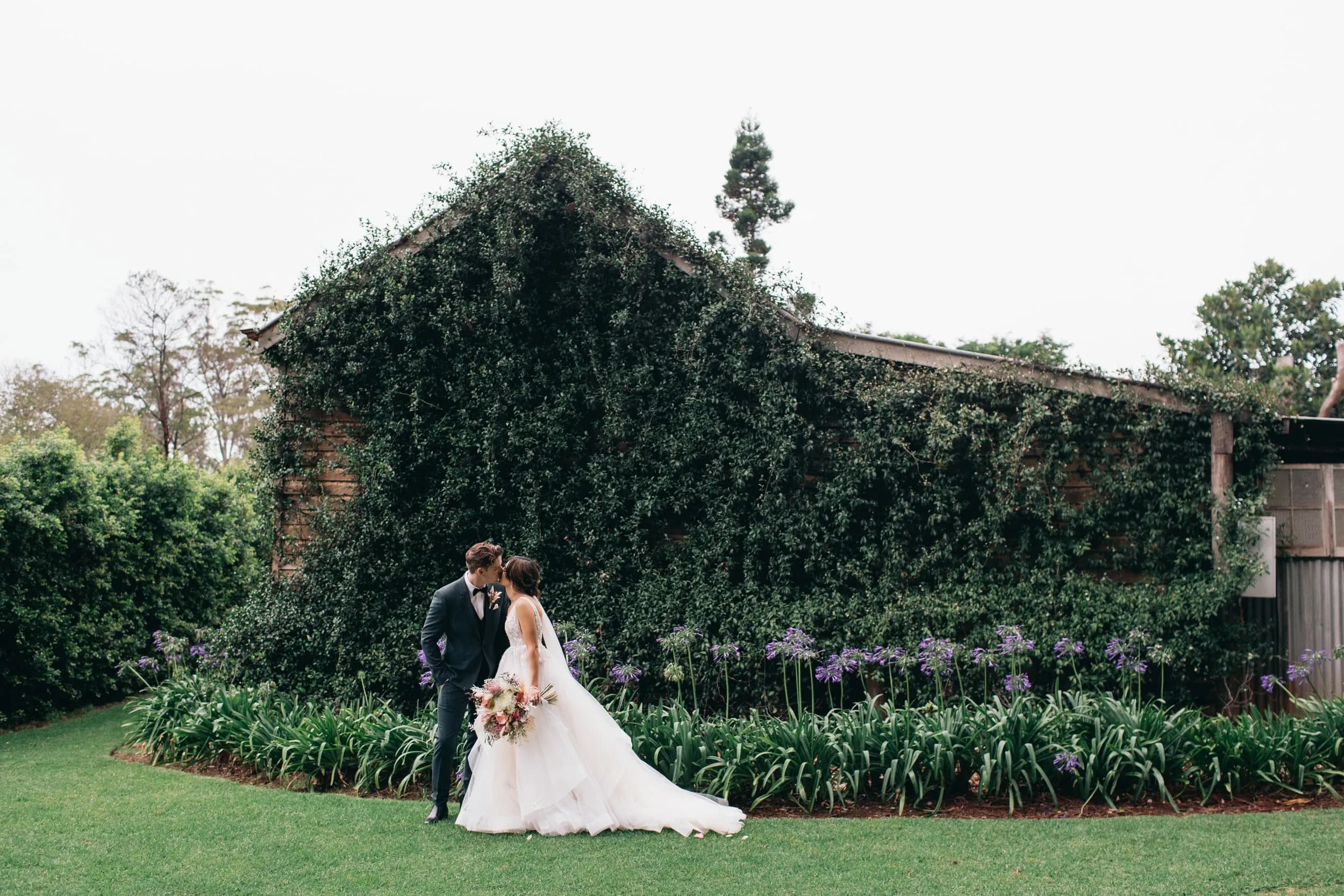 A bride and groom stand close together, lovingly gazing at each other in front of a vine-covered building. The bride is in a white wedding dress holding a floral bouquet, and the groom is in a dark suit. Surrounding them are lush green plants and purple flowers.