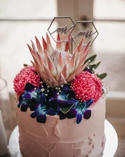 A pink frosted cake topped with vibrant flowers and greenery. At the center is a large protea flower, surrounded by pink dahlias and purple orchids. Above the flowers, two hexagon topper signs read "Mr" and "Mrs." The cake has a textured rustic finish.