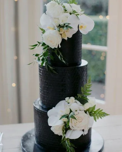 A three-tiered black wedding cake is elegantly decorated with clusters of white flowers and green leaves. The cake features intricate textures on each tier, and the background shows softly lit windows and greenery outside.