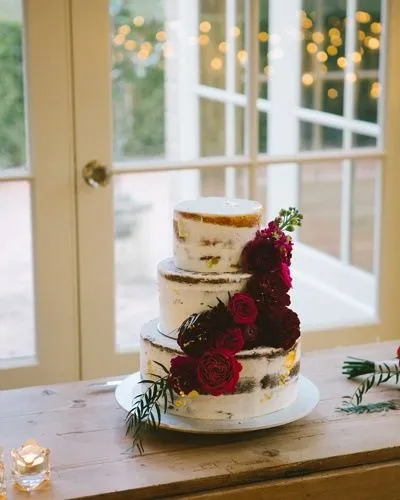 A three-tier naked wedding cake adorned with dark red roses and greenery sits on a wooden table in front of a glass door with string lights in the background.