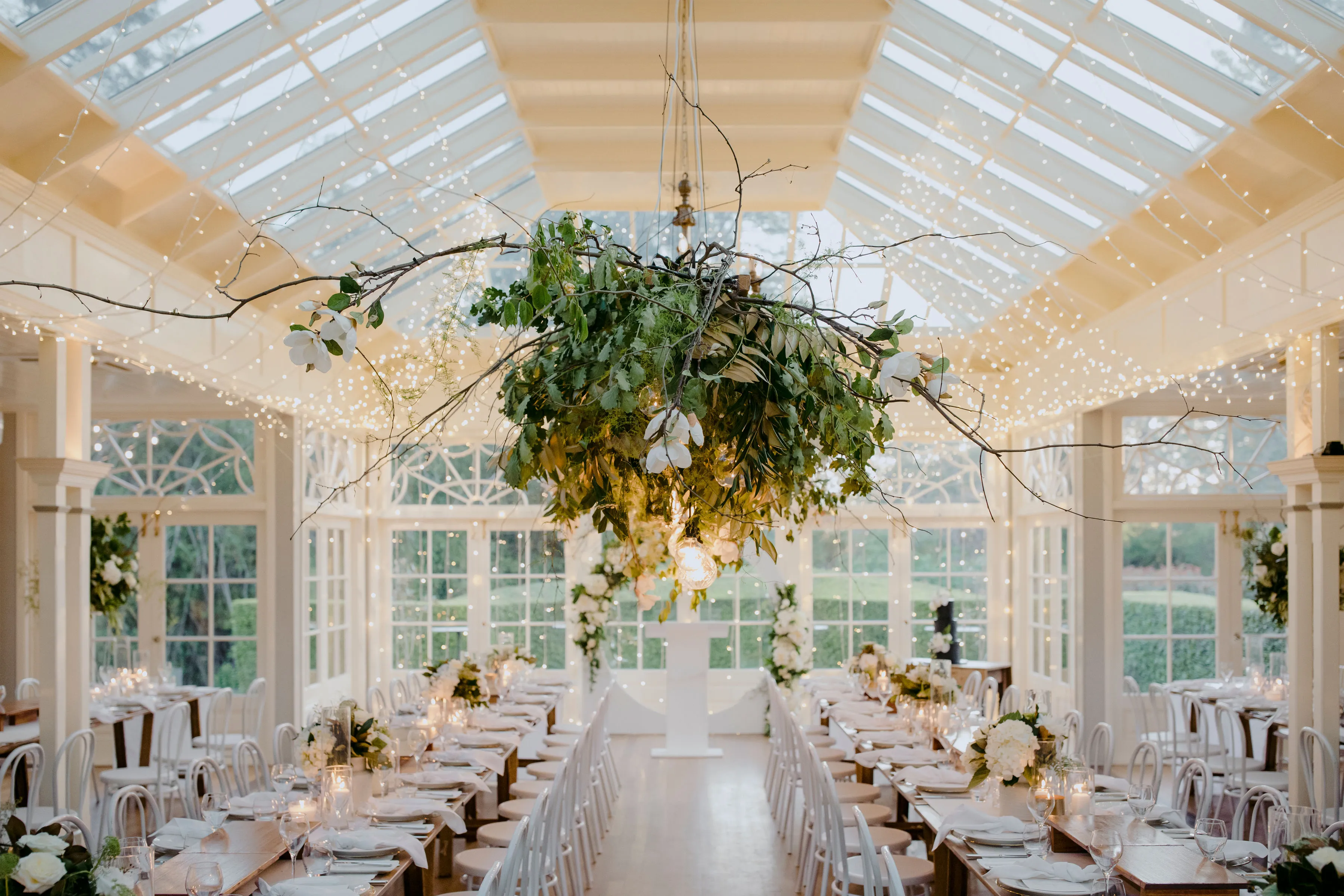 A beautifully decorated indoor wedding venue features a high glass ceiling adorned with strings of fairy lights. Elegant wooden tables are arranged in rows, each decorated with white flowers and greenery. A large, floral chandelier hangs centrally, creating a focal point.