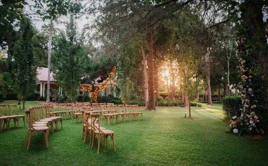 An outdoor wedding setup in a lush green garden with rows of wooden chairs arranged on the grass. Twinkling string lights are draped around a tree, and the sun is setting in the background, casting a warm glow through the trees creating a serene atmosphere.
