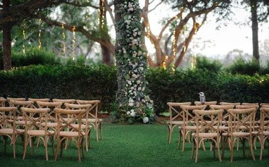A serene outdoor wedding setup with rows of wooden chairs arranged on a green lawn facing an aisle, adorned with floral decorations. String lights are hanging from trees in the background, creating a warm, inviting atmosphere in a lush garden setting.