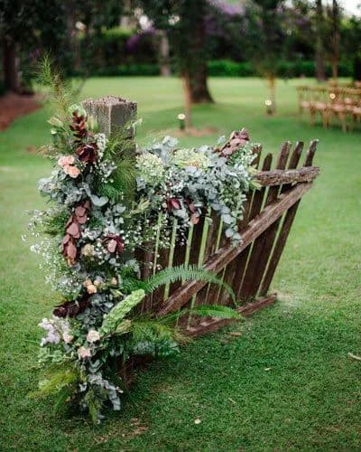 A rustic wooden fence adorned with lush green foliage and flowers, set against a grassy outdoor backdrop. The fence appears weathered, adding to the natural, earthy charm of the scene, which could be part of an outdoor event or wedding setup.