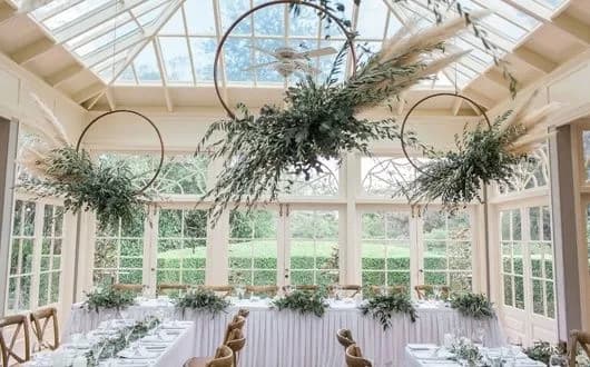 Elegant greenhouse wedding setup with large glass windows and ceiling, natural light, and greenery decorating the space. Circular tables with white tablecloths and green foliage centerpieces fill the room. Suspended floral arrangements add a touch of nature.