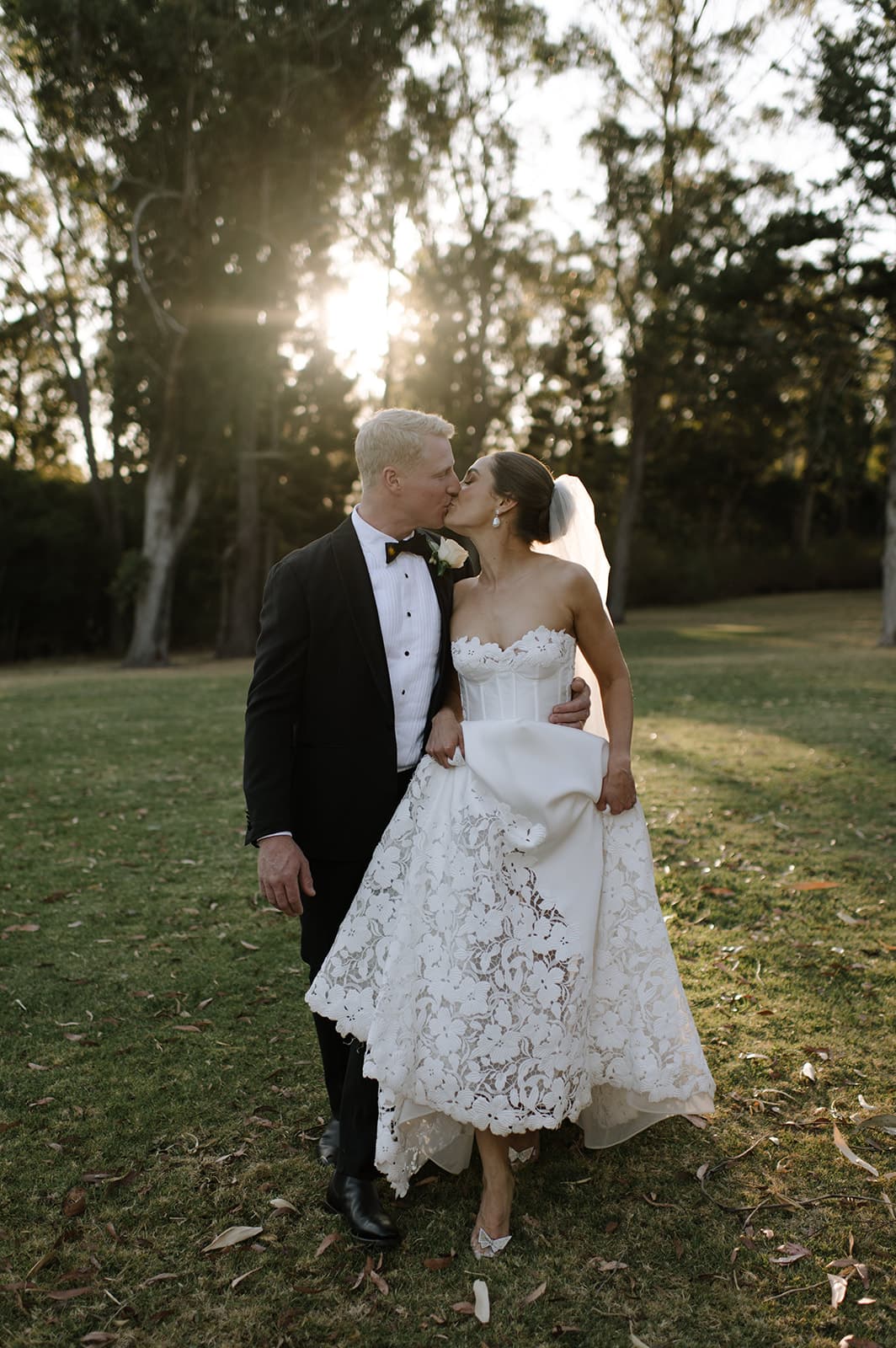A newlywed couple shares a kiss outdoors during sunset. The groom, in a black tuxedo and bow tie, holds the bride, who wears an elegant white lace wedding gown and veil. They stand on a grassy area with tall trees in the background, bathed in warm sunlight.