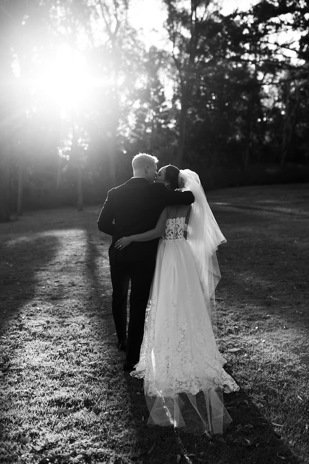 A newlywed couple is walking away down a grassy path with their arms around each other. The groom is dressed in a dark suit and the bride is wearing a strapless gown with a long veil. The sun is shining through the trees in the background, creating a serene atmosphere.