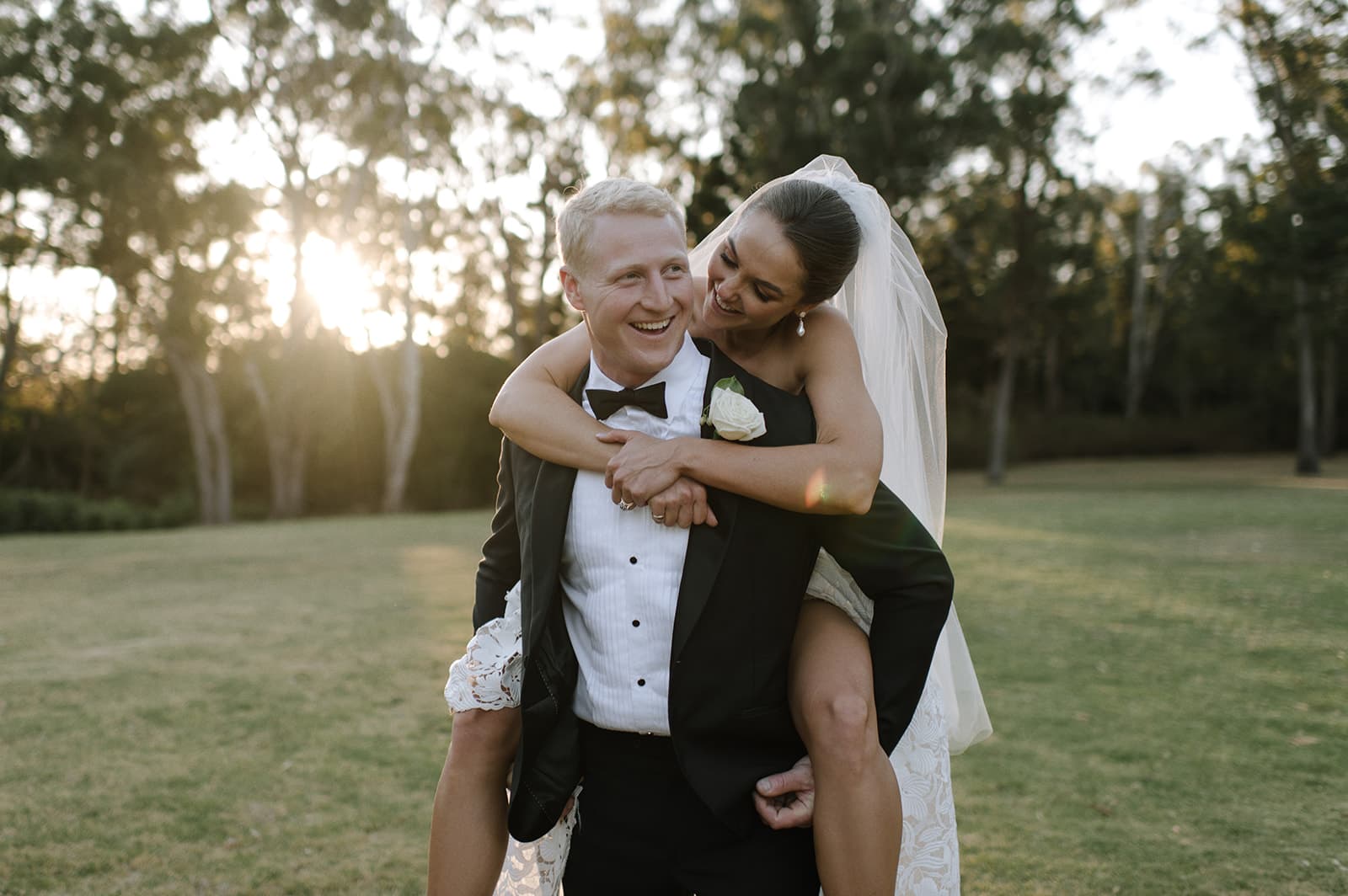 A groom in a black tuxedo carrying his bride, who is in a white wedding dress and veil, on his back. They are both laughing and standing on a grassy field with trees in the background. The setting sun creates a warm glow around them.