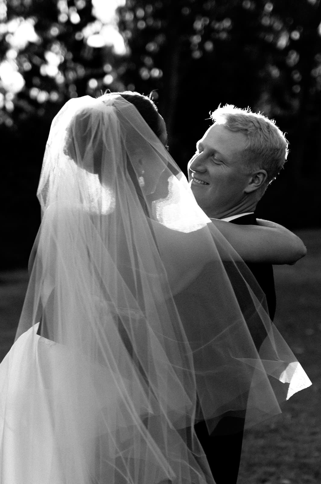 A newlywed couple shares a joyful moment, with the groom gazing lovingly at the bride. The bride's veil and dress are beautifully illuminated in the soft, natural light. The background is softly blurred, emphasizing the couple’s happiness on their special day.