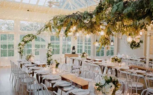 A beautifully decorated wedding reception with white tables and chairs, adorned with greenery, flowers, and hanging lights. The setting is bright with large windows and a glass ceiling, allowing natural light to fill the room.