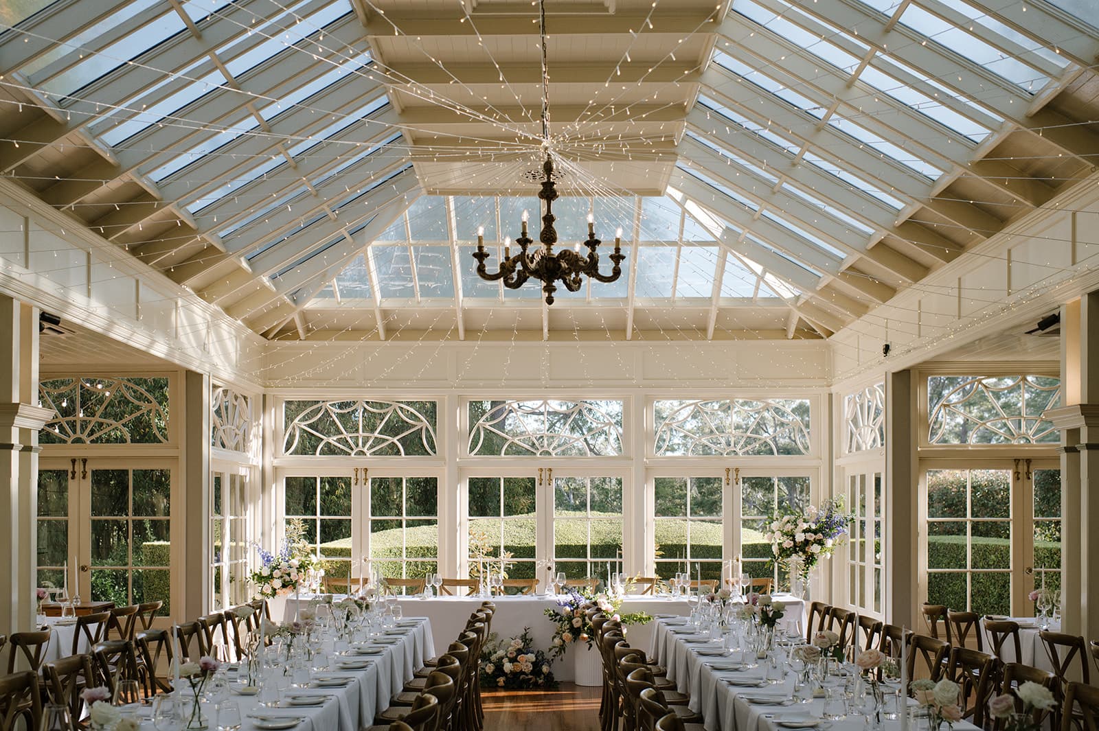 A beautifully set dining area in a sunlit conservatory with glass walls and ceiling. Long tables are adorned with elegant white tablecloths, floral arrangements, and fine dining ware. A vintage chandelier hangs from the ceiling, adding a touch of sophistication.