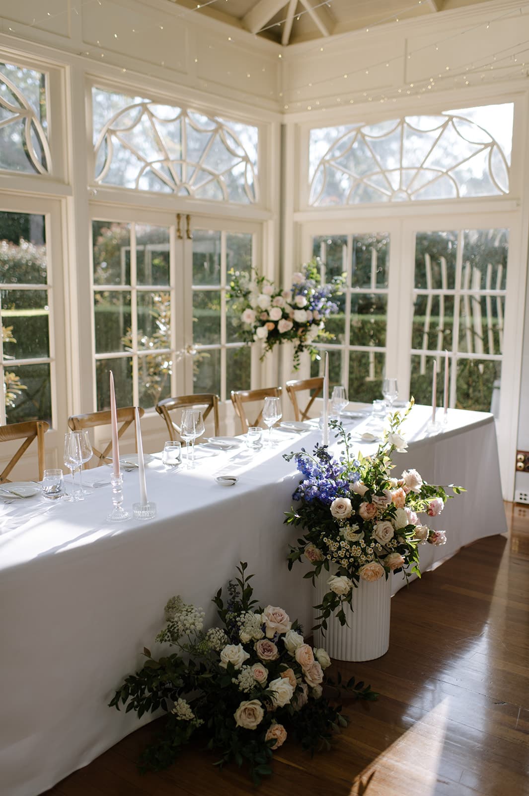 A beautifully decorated wedding reception table set in a sunlit room with large windows. The table is adorned with white linens, glassware, and tall candles, with elegant floral arrangements featuring blue, white, and pink flowers both on and beside the table.