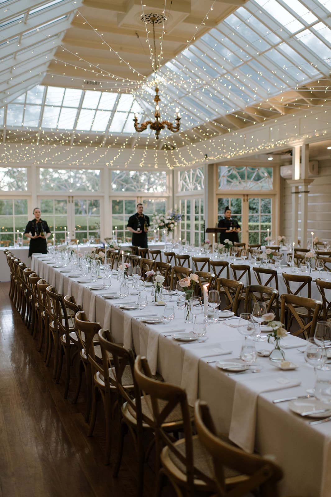 A beautifully set, long dining table is prepared for an event in a spacious, well-lit room with large windows. The table is adorned with white tablecloths, glassware, cutlery, and simple floral centerpieces. String lights hang from the ceiling, creating a warm ambiance.