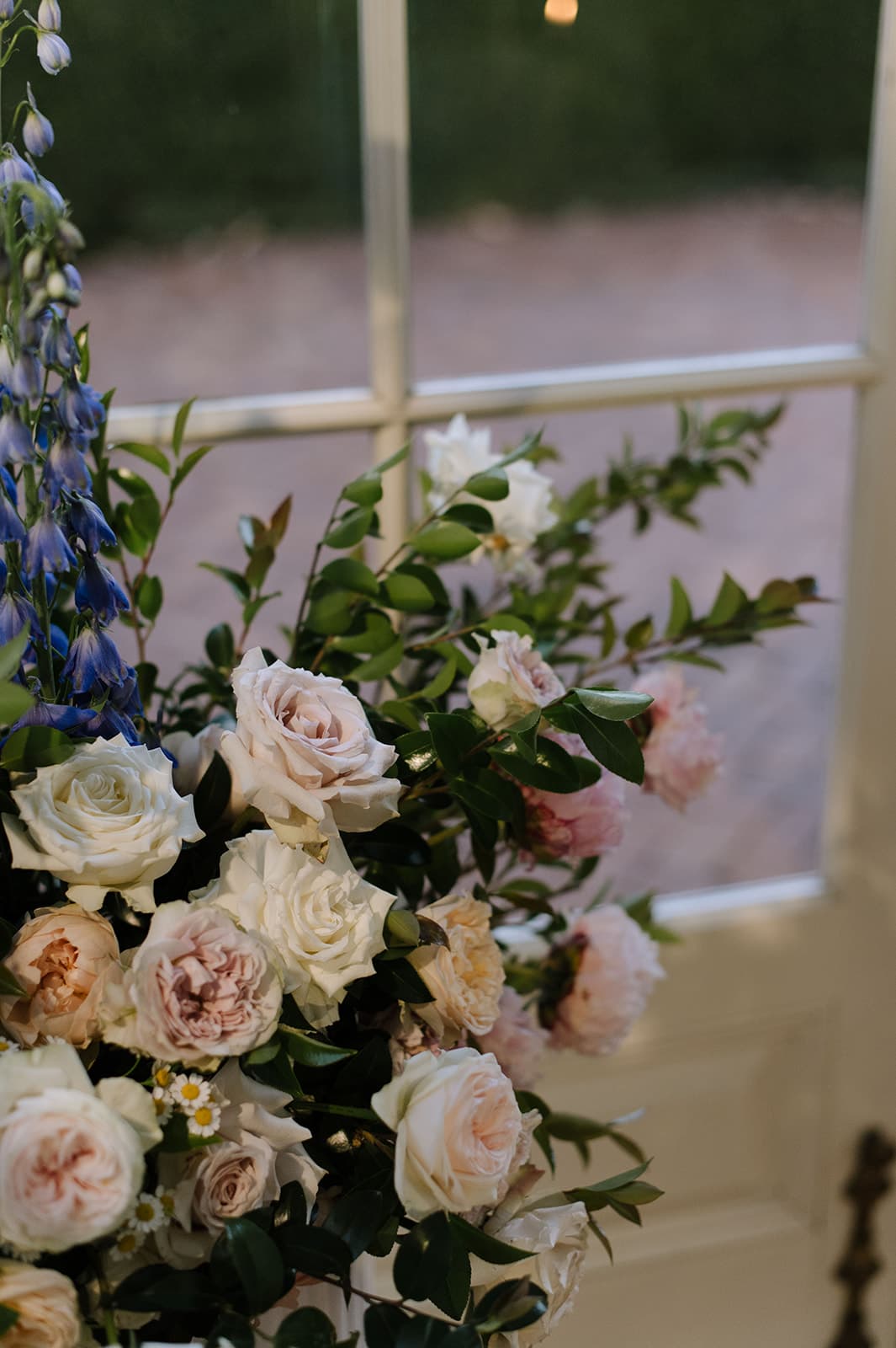 A floral arrangement with pastel roses, peonies, and greenery is positioned in front of a glass door. The soft pink, white, and peach flowers are complemented by green leaves and a few tall, blue flowers, with a brick patio visible outside through the glass.
