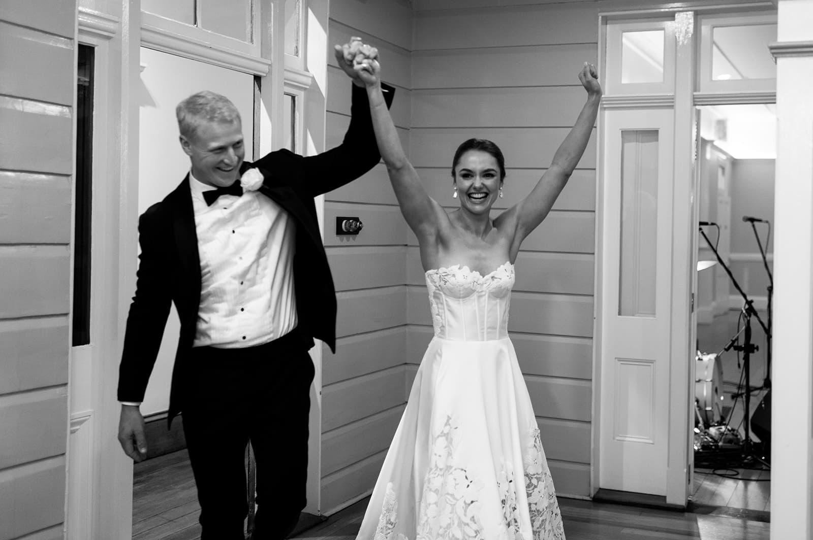 A newly married couple, dressed in formal attire, enters a room holding hands up high in celebration. The groom wears a black suit with a white shirt and bow tie, and the bride wears a strapless white gown. Both are smiling and appear joyful. The photo is in black and white.