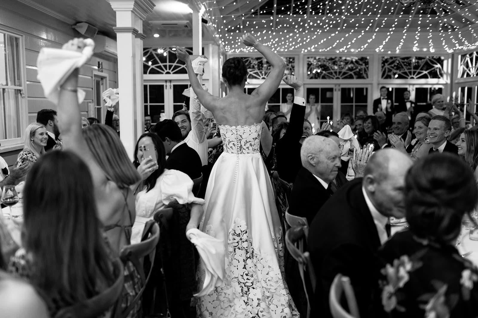 A bride in a strapless gown with a lace overlay stands with her back to the camera, lifting her arms and holding a napkin. She is surrounded by guests at a wedding reception, who are seated at long tables and also waving napkins in a festive, well-lit room.