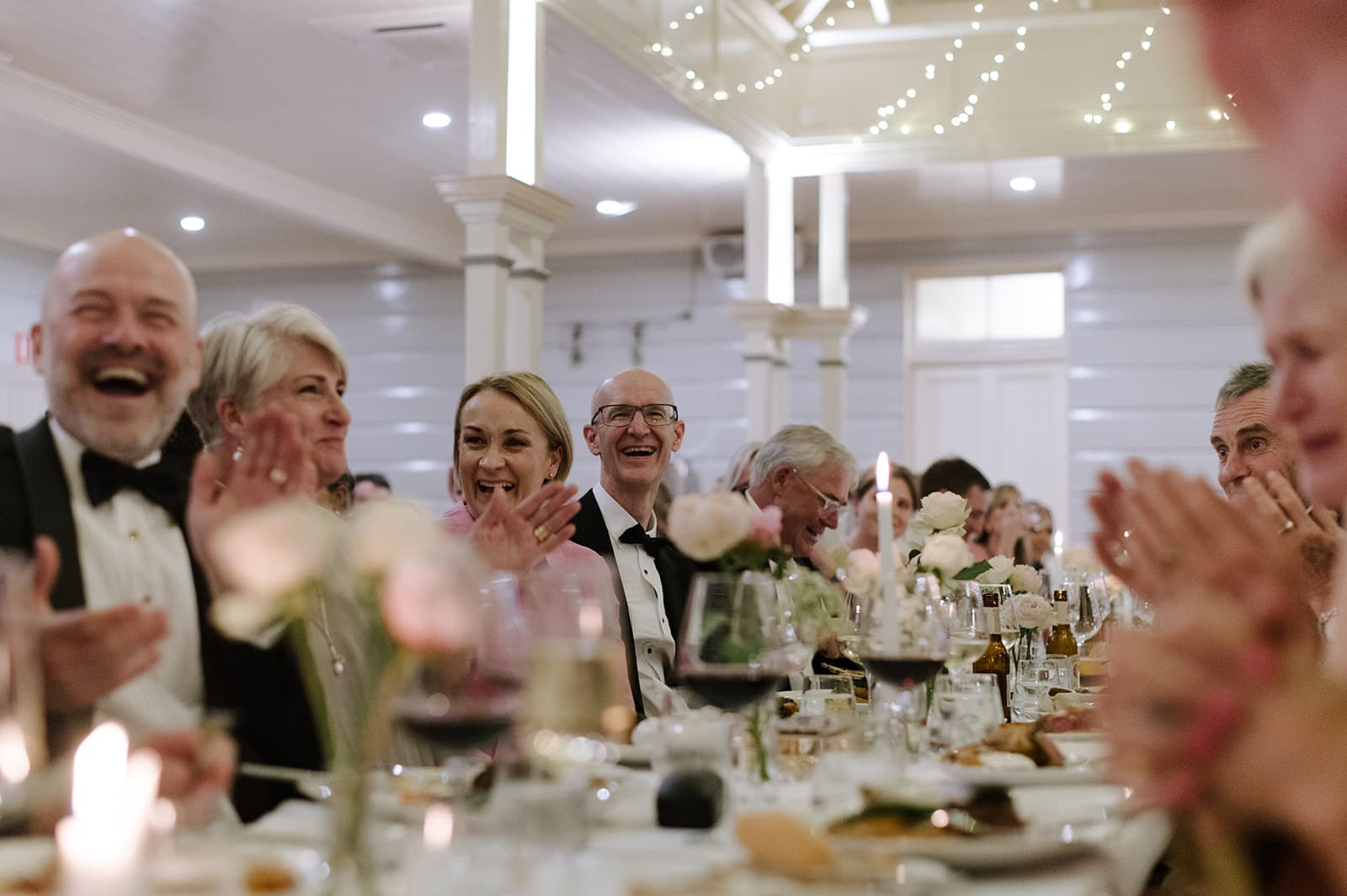 People dressed in formal attire are sitting at a long table, smiling, laughing, and clapping. The table is set with elegant dinnerware, candles, flowers, and wine glasses. Twinkling string lights are hung from the ceiling, creating a festive atmosphere.