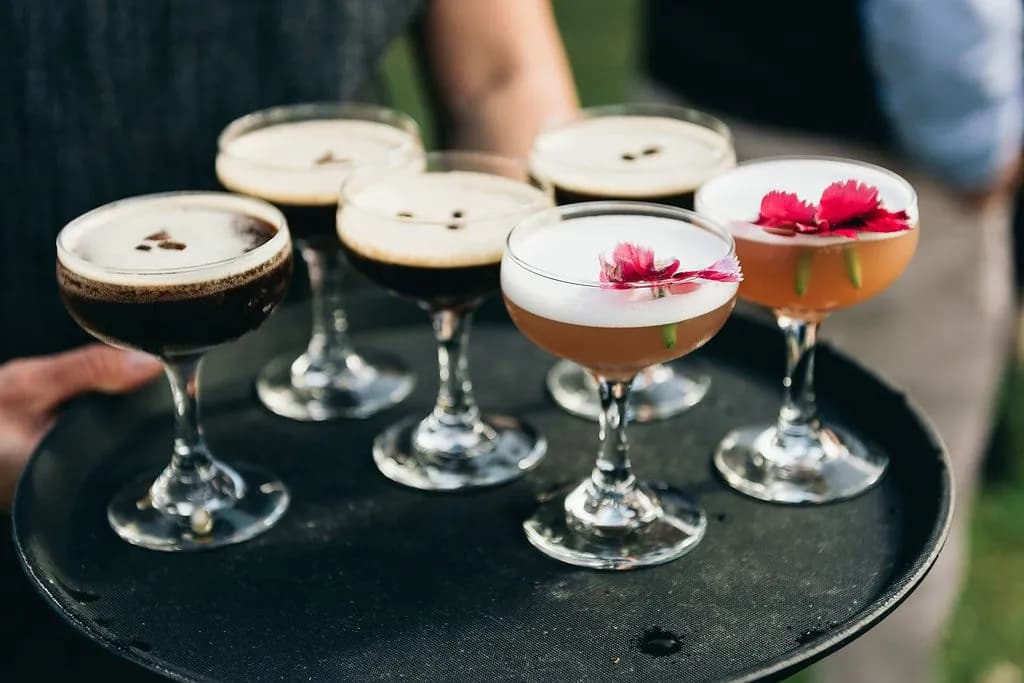 A person holds a black tray with seven cocktails in coupe glasses. Four drinks are dark with foam on top, garnished with three coffee beans each. The other three drinks are lighter in color, topped with foam and decorated with bright pink flowers.