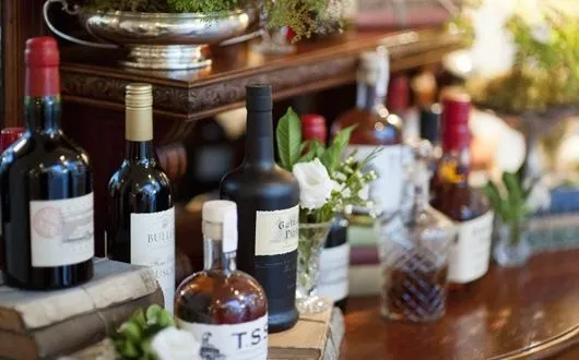 A collection of various wine and liquor bottles displayed on a polished wooden surface, some elevated on books. Greenery and white flowers are arranged among the bottles, creating an elegant and inviting setting.