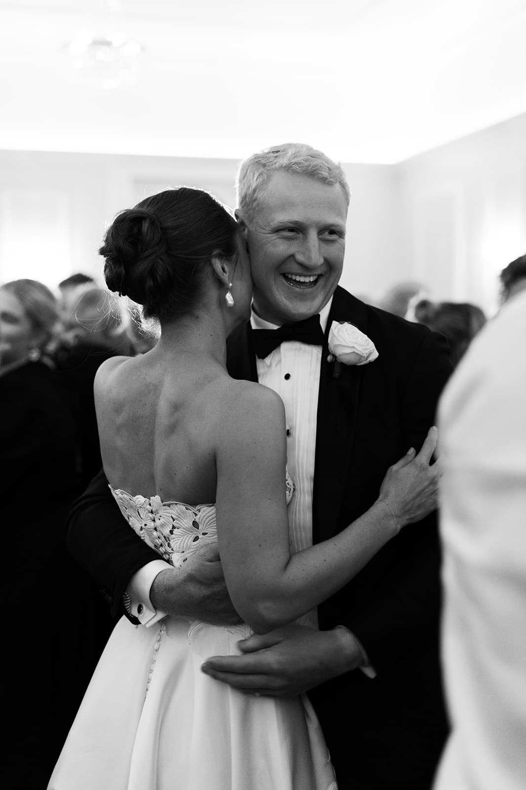 A groom in a tuxedo with a bow tie and a boutonniere smiles while hugging a bride in an off-the-shoulder wedding dress. The bride has her back to the camera, and they are surrounded by other people in formal attire.