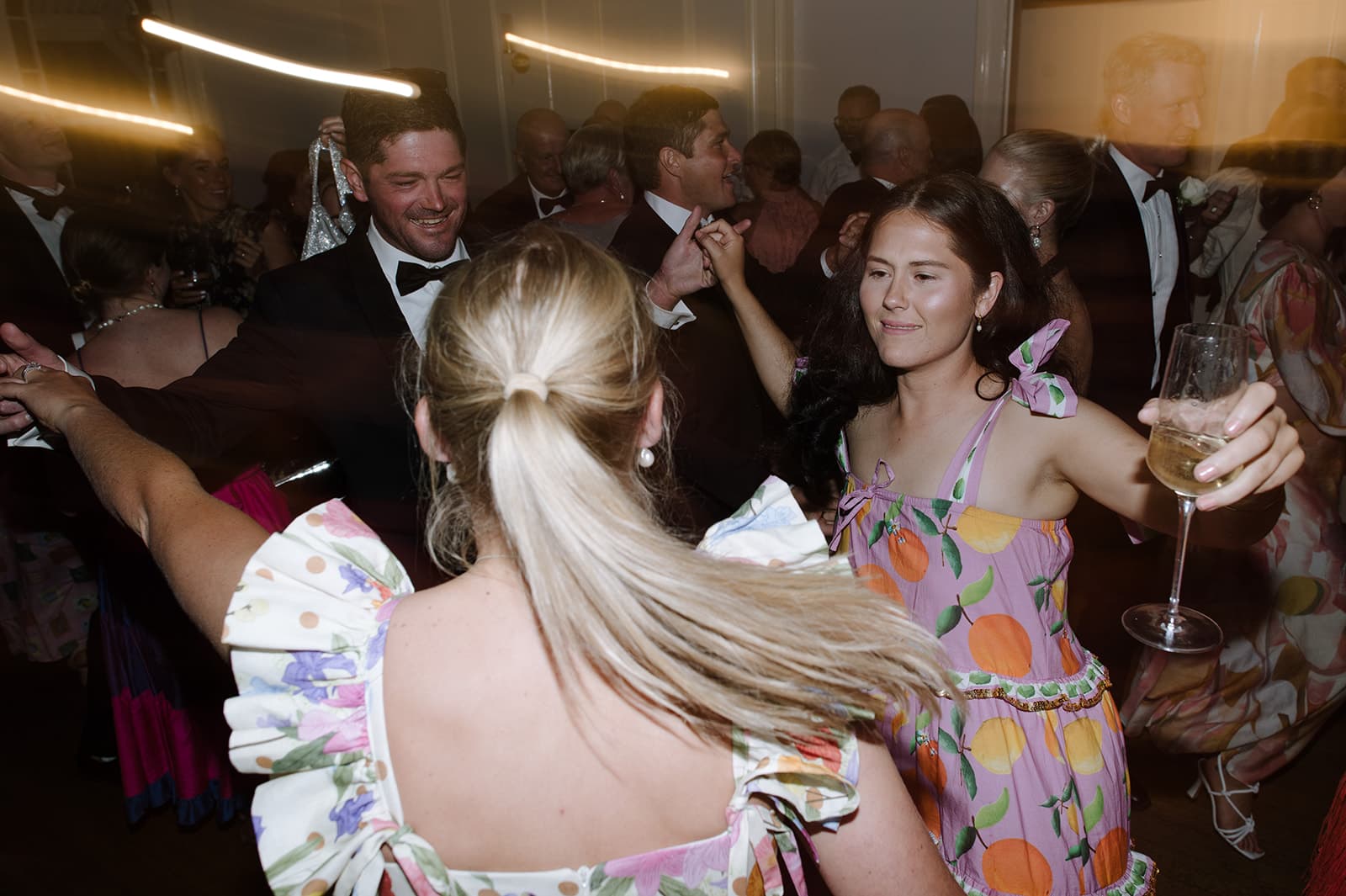 A lively party scene with people dancing. A woman in a colorful dress holds a wine glass, smiling and dancing with another woman in a floral dress with a ponytail. A man in a suit and bow tie dances nearby. The room is filled with other people enjoying the event.