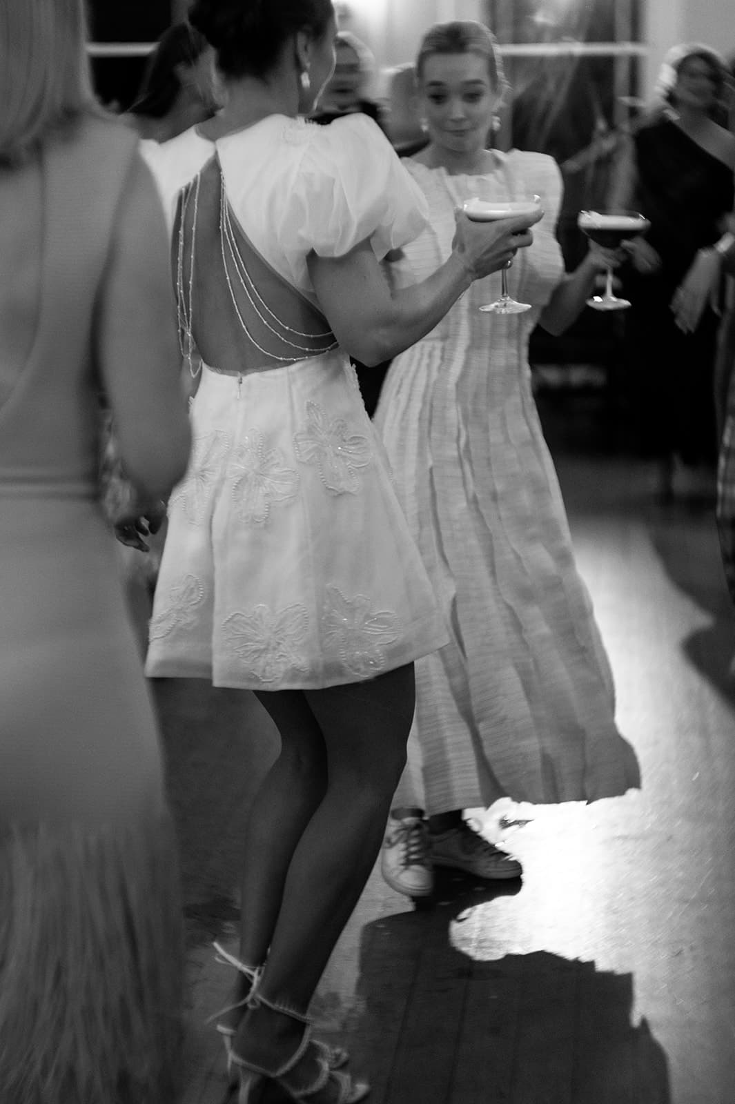 Black and white photo of two women in dresses dancing at an indoor event. One woman holds a cocktail glass in one hand while touching the other woman's arm. Both are smiling and seem engaged in a pleasant conversation. Others and a lit window are visible in the background.