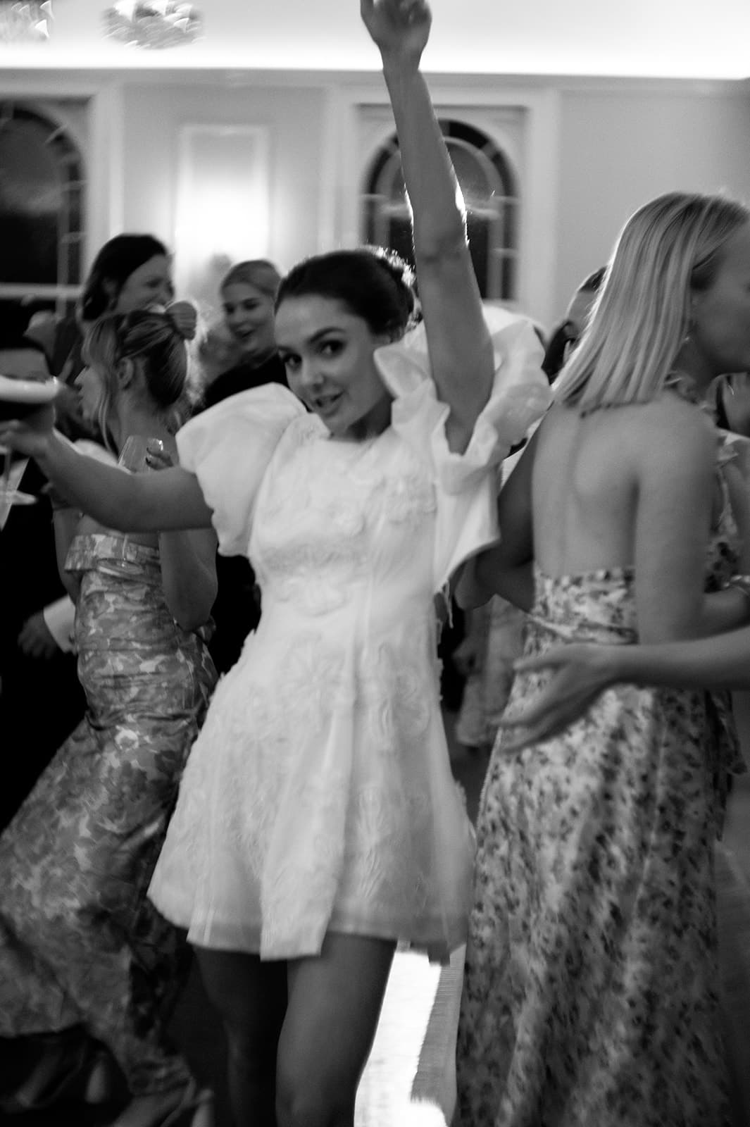 A joyful woman in a short white dress dances with one arm raised at a lively indoor event. Other people in elegant attire are dancing and socializing around her. The photo is in black and white, creating a timeless, celebratory atmosphere.